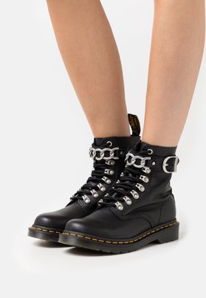 Bottes cheville en cuir noir avec détails de chaîne et boucle argentées portées sur des jambes nues sur fond blanc.