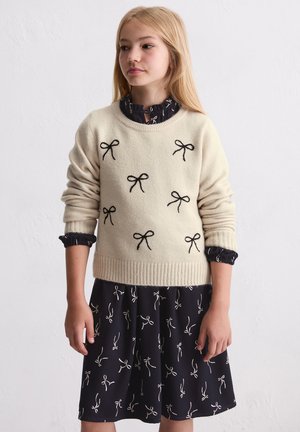 Crèmesweater met zwarte strikbroderie, geribbelde zoom, gedragen over een donkere jurk met een vergelijkbaar strikpatroon. Gladde textuur, ontspannen pasvorm.