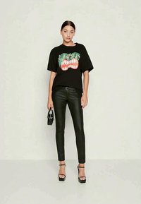 Melna grafiska t-krekla ar zaļu un sarkanu tekstu un zobu dizainu, saskaņota ar melniem pieguļošiem biksēm un siksnām augstpapēžu sandālēm.