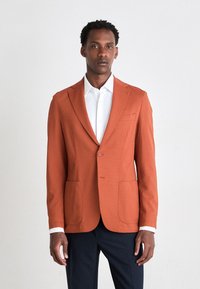 Orange texturerad blazer med två framfickor, enkel knappstängning och slagluckor. Bärs över en vit skjorta med marinblå byxor.