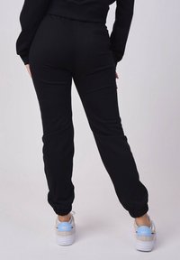 Osoba ubrana w czarne dresowe spodnie i białe tenisówki z jasnoniebieskimi akcentami, stojąca plecami do widza na jednolitym tle.