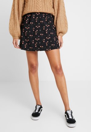 Czarna spódnica w kwiaty o rozkloszowanym kroju, ozdobiona małymi różowymi kwiatami. Noszona z beżowym swetrem z dzianiny i czarnymi trampkami.