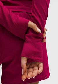 Langsleeve shirt en shorts in een levendige fuchsia kleur, met ribbelstructuur, manchetdetails en een ontspannen pasvorm. Een hand is zichtbaar en grijpt de mouw.