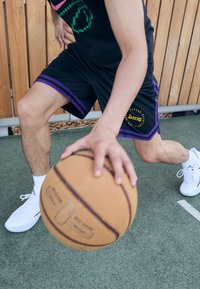 Čierne basketbalové šortky s fialovými akcentmi a logom Lakers, v kombinácii s bielymi topánkami. Osoba dribluje hnedým basketbalom na ihrisku.