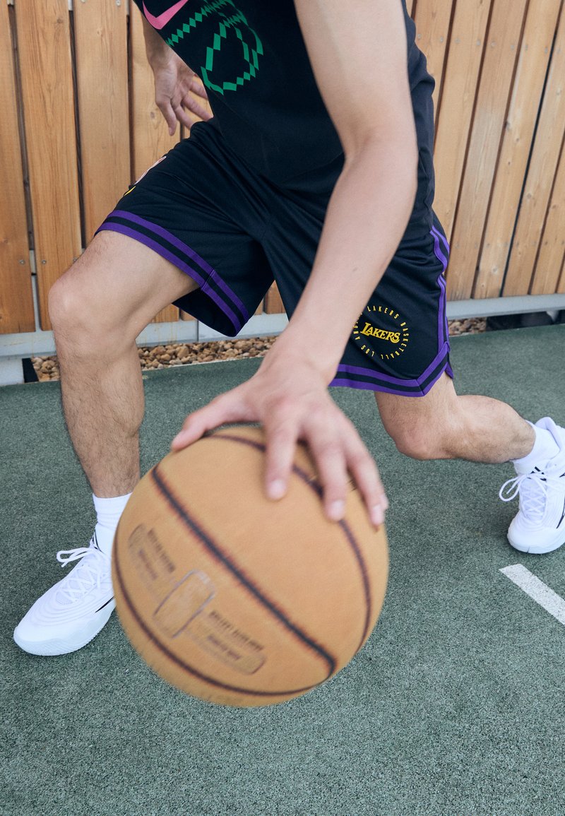 Čierne basketbalové šortky s fialovými akcentmi a logom Lakers, v kombinácii s bielymi topánkami. Osoba dribluje hnedým basketbalom na ihrisku.