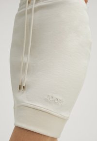 Beige Baumwollshorts mit einem Kordelzug, gerippten Bündchen und einem dezenten Logo-Stick auf dem Bein. Glatte Textur insgesamt.