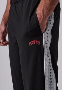 Άνδρας ντυμένος με μαύρο παντελόνι που έχει γκρι ρίγα στο πλάι με μαύρες σιλουέτες και κόκκινο λογότυπο, με το χέρι στην τσέπη, μπροστά σε απλό φόντο.