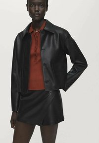 Zwart leren cropped jack met een kraag, knoopsluiting aan de voorkant en twee zakken, gedragen over een geribde rode top en een zwarte mini-jurk.