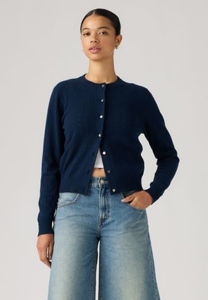 Femme portant un cardigan bleu foncé boutonné sur un top blanc court avec un jean taille haute bleu clair, debout avec une main dans la poche.