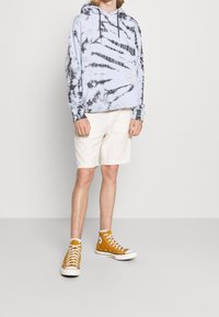 Persona vestida con una sudadera de teñido anudado en blanco y negro, pantalones cortos crema y zapatillas altas mostaza, de pie contra un fondo gris liso.