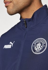 Mornarska zip-up jakna z logotipom Puma in grbom Manchester City, izdelana iz gladkega materiala z visokim ovratnikom in subtilnimi šivi.