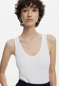 Ung person med kort lockigt hår bär en vit ärmlös topp med rundad hals mot en enfärgad ljus bakgrund.