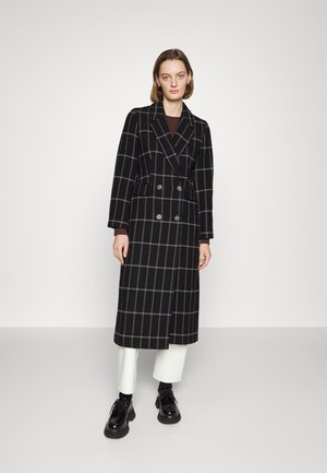 Black plaid coat with a double-breasted design, featuring large buttons and a long silhouette. Worn over white trousers and black shoes.