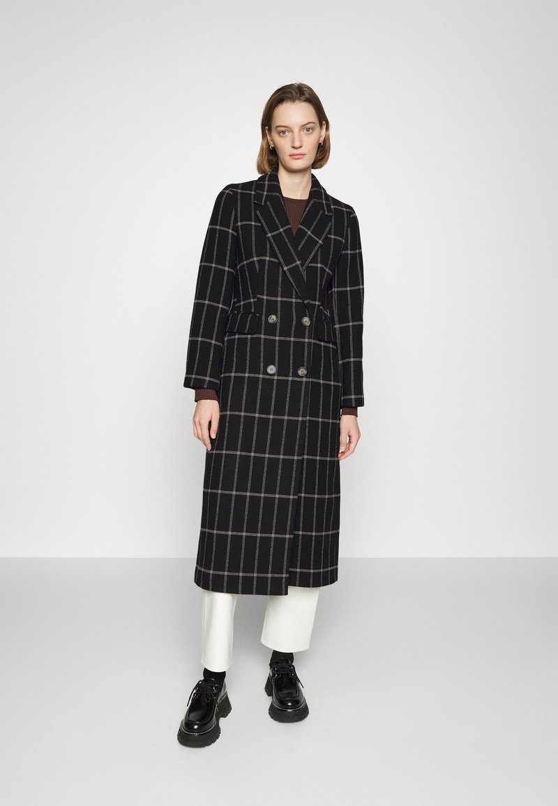 Cappotto a quadri neri con design doppiopetto, caratterizzato da grandi bottoni e una silhouette lunga. Indossato sopra pantaloni bianchi e scarpe nere.
