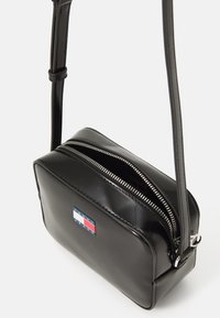 Bolso cruzado de charol negro con forma rectangular, cierre de cremallera y un parche con logotipo que presenta franjas rojas, blancas y azules.