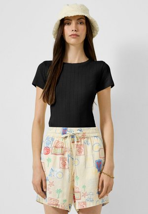 Femme portant un haut noir moulant à manches courtes, un short crème avec des imprimés colorés sur le thème du voyage, un chapeau bob beige et un bracelet en or.