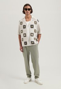 Korte mouwen, witte haakshirts met multikleurige vierkante motieven, gecombineerd met lichtgroene broek en grijze sneakers. Zonnebril maakt de look compleet.