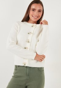 Cárdigan de punto blanco con manga larga, que presenta botones decorativos en crema y verde. Textura suave, diseño corto y puños acanalados.