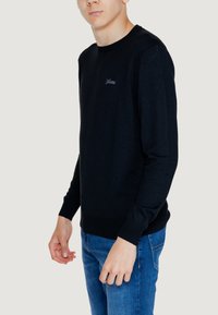 Maglione nero a maglia con colletto girocollo, polsini e orlo a coste, caratterizzato da un piccolo logo ricamato sul petto, abbinato a jeans blu.