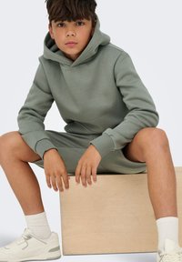 Hætte sweatshirt i afdæmpet grøn, lavet af blødt stof. Har ribbede ærmer og en løs pasform, sammen med hvide sneakers og ankelsokker.
