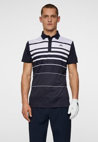 Marinblå och vit randig polo med krage, korta ärmar och en logotyp på bröstet. Bärs med mörka byxor och en vit golfhandske.