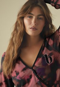 Patterned long-sleeve top featuring pink and black hues, ribbed texture, deep V-neck, and a contrasting embroidered letter design.