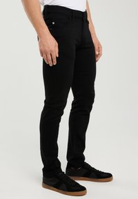 Männliches Model trägt schwarze Slim-Fit-Jeans und schwarze Sneaker mit Gummisohlen, steht vor einem schlichten hellen Hintergrund.