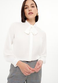 Blusa blanca con un lazo en el cuello, con mangas largas y una textura de tela suave, combinada con pantalones de cuadros grises.