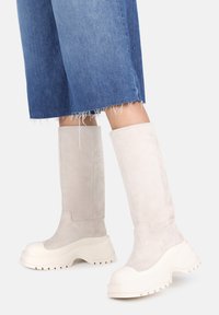 Knee-high šviesiai rudos zomšinės botai su stambiu baltu guminiais padu ir apvaliu nosiu, derinami su trumpomis mėlynomis džinsinėmis kelnėmis, turinčiomis nutrintą kraštą.