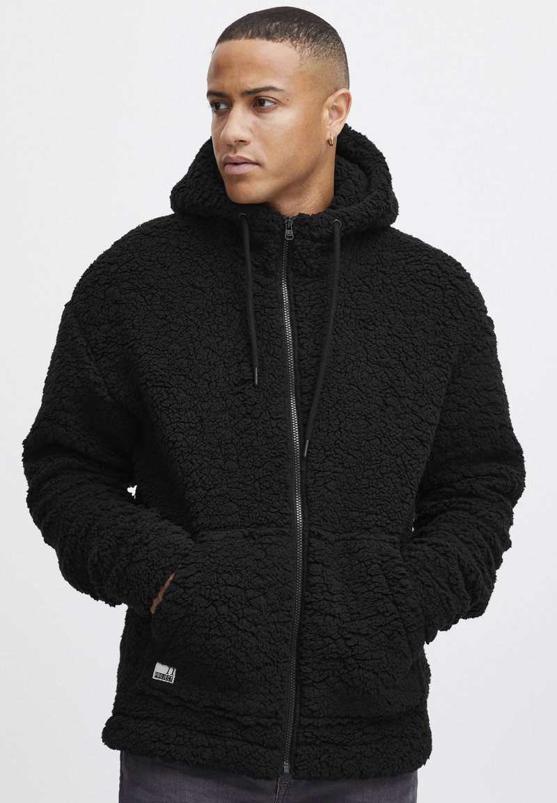 Black fleece zip-up hoodie with a textured pattern, drawstring hood, and front pockets. Features a logo patch on the lower hem.