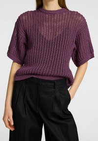 Woman wearing a loose, short-sleeve purple knit sweater over a black top, paired with high-waist black pleated trousers, one hand in pocket.