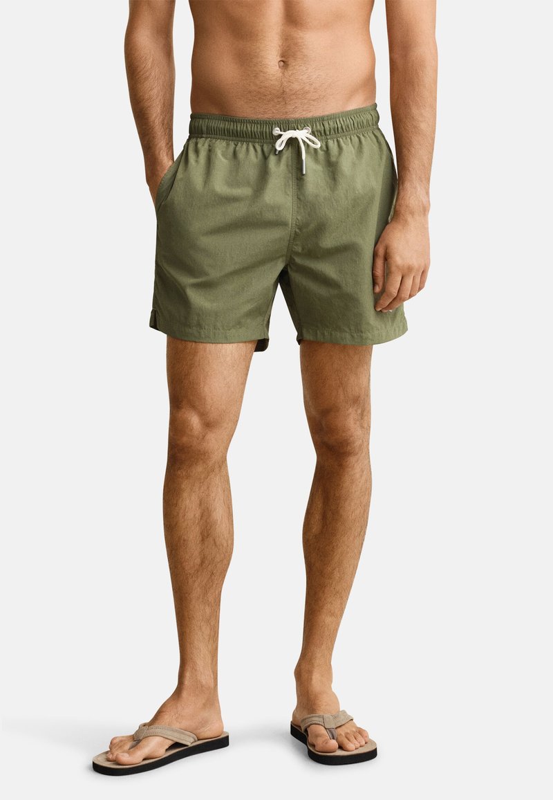 Homme portant un short de bain vert olive avec un cordon blanc et des tongs beiges, debout devant un fond clair uni.
