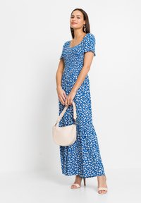 Abito floreale blu con maniche a sbuffo corte, scollatura quadrata e gonna svasata. La modella tiene una borsa color crema con una tracolla intrecciata.