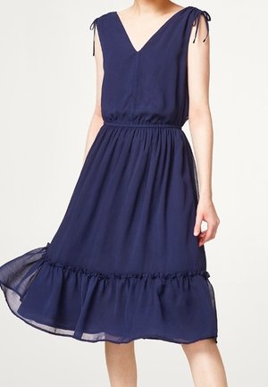 Granatowa sukienka midi bez rękawów z dekoltem w serek, marszczonym stanem, wiązaniami na ramionach i falbaną na dole, noszona przez stojącą osobę.