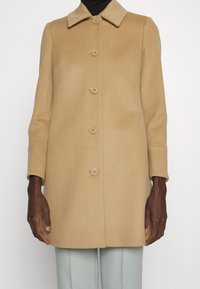 Manteau en mélange de laine couleur tan avec une silhouette structurée, un col pointu et six boutons sur le devant. Manches longues et texture lisse.