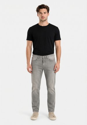 Giovane uomo con capelli castani che indossa una maglietta nera a maniche corte, jeans grigi chiari e sneakers beige, in piedi contro uno sfondo bianco uniforme.