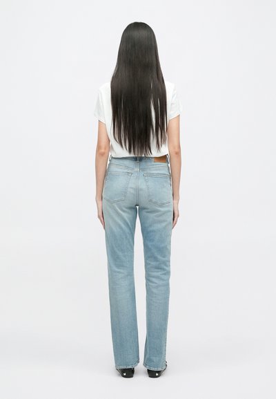 Jeans de cintura alta azul claro com ligeiro alargamento, apresentando quatro bolsos traseiros e uma etiqueta de couro bege. Usados com uma t-shirt branca justa.