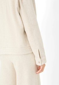 Helle beige, strukturierten Stoffjacke mit seitlichen Schlitzen und geknöpften Manschetten. Das Design zeichnet sich durch sichtbare Nähte entlang des Rückens und des Saums aus.
