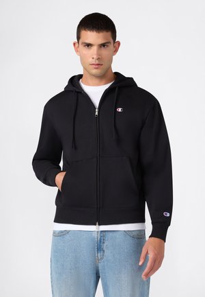 Junger Mann trägt schwarzen Kapuzenpullover mit Reißverschluss, Logo auf Brust und Ärmel, weißes Hemd darunter und hellblaue Jeans, steht vor weißem Hintergrund.