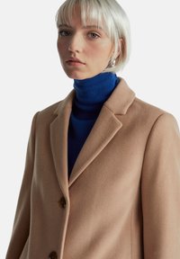 Světle hnědý vlněný kabát se strukturovaným límcem a dvěma knoflíky, vrstvený přes modrý žebrovaný rolák. Hladká textura, přizpůsobený střih.