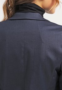 Marinblå blazer i texturerat tyg, med slagkrage och bakre sömmar. En tunn svart polotröja syns under.