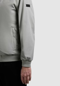 Lys grå jakkeærme med elastisk manchet, sidelomme og KHUJO-mærkpatch på den øvre arm, vist på en stående person.