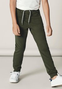 Pantalons de jogging vert olive avec une taille élastique, dotés d'un cordon de serrage et de poignets côtelés, portés avec des baskets blanches. Pas de motifs visibles.