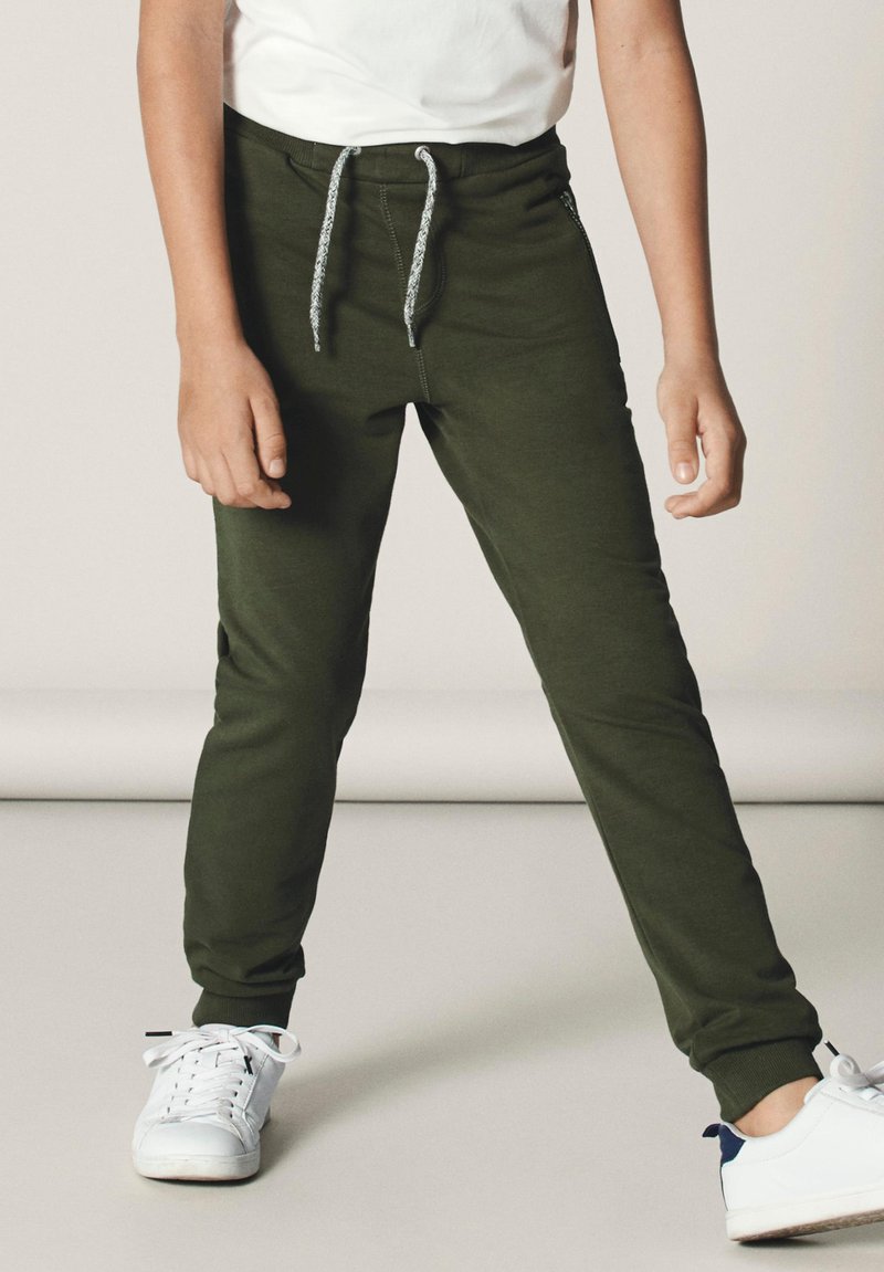 Pantalons de jogging vert olive avec une taille élastique, dotés d'un cordon de serrage et de poignets côtelés, portés avec des baskets blanches. Pas de motifs visibles.