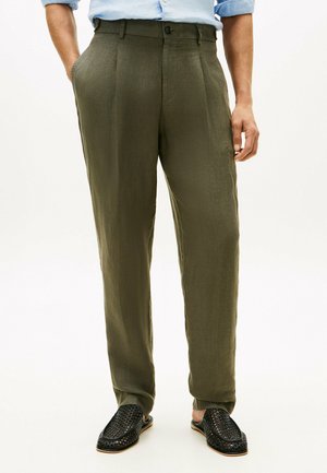 Mann trägt olivgrüne gerade geschnittene Hosen, schwarze gewebte Slipper und ein hellblaues Hemd mit hochgekrempelten Ärmeln.