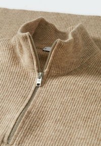 Pull en maille côtelée beige avec une fermeture éclair métallique à un quart et un col haut replié.
