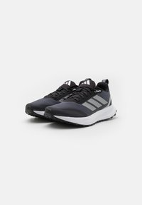 Schwarze und graue Sportschuhe mit einer strukturierten Oberseite, kontrastierender weißer Mittelsohle, reflektierenden Akzenten und einem gemusterten Schnürsystem.