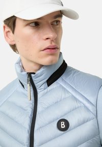 Hellblaue gesteppt wattierte Weste mit hohem Kragen, schwarzen Mesh-Akzenten und einem runden Logo-Patch. Reißverschluss mit metallischem Zippergriff.