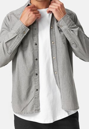 Mann, der ein hellgraues Jeanshemd über einem weißen T-Shirt zuknöpft, trägt schwarze Hosen, sichtbar vom Hals bis zu den Hüften.