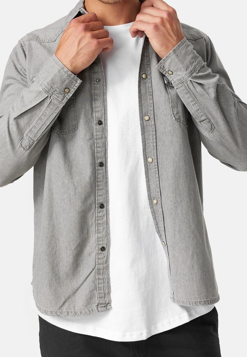 Homme boutonnant une chemise en denim gris clair par-dessus un t-shirt blanc, portant un pantalon noir, visible du cou aux hanches.